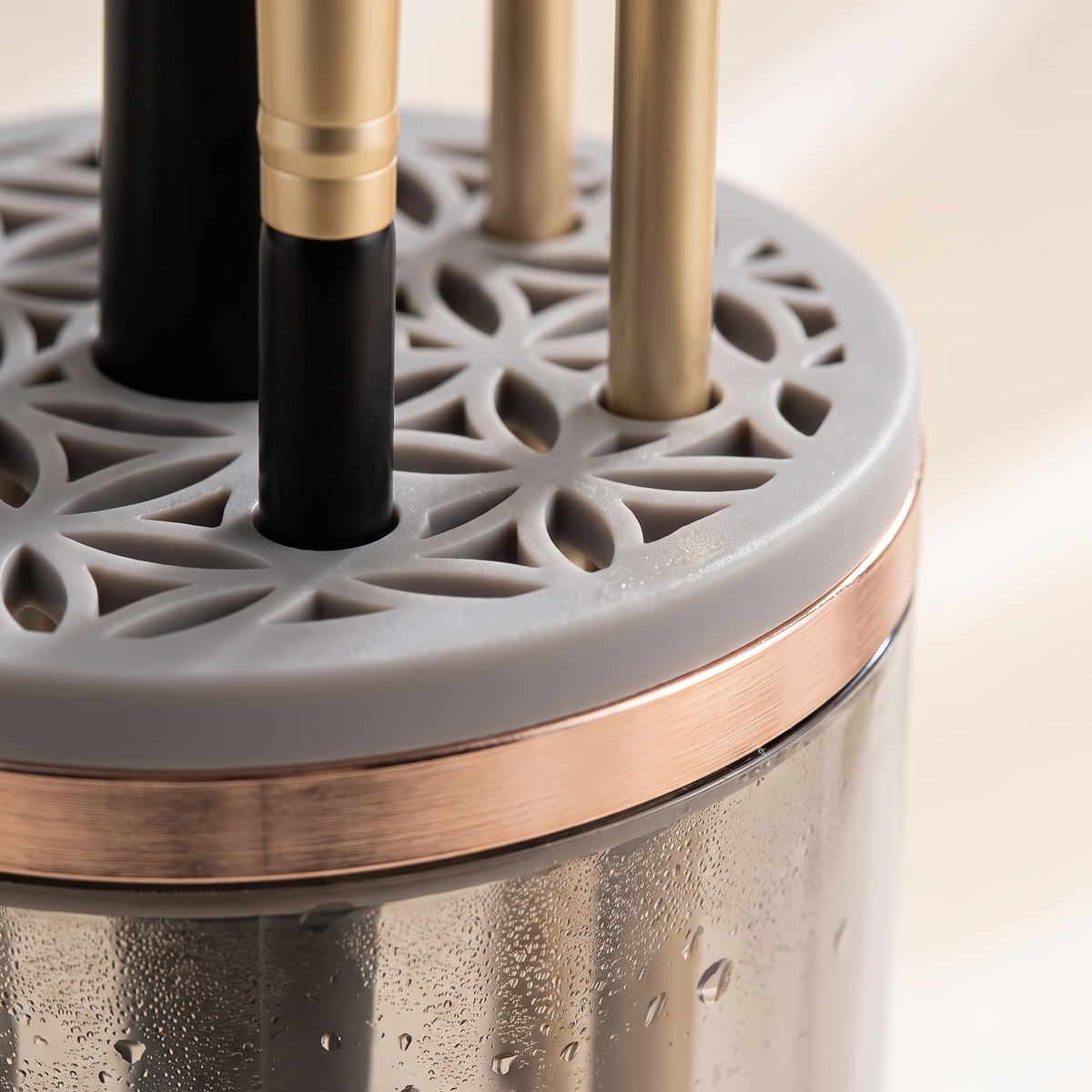 Close-up of a makeup brush holder with a gray perforated lid, rose gold trim, and several brushes inserted, including black and gold-handled brushes.